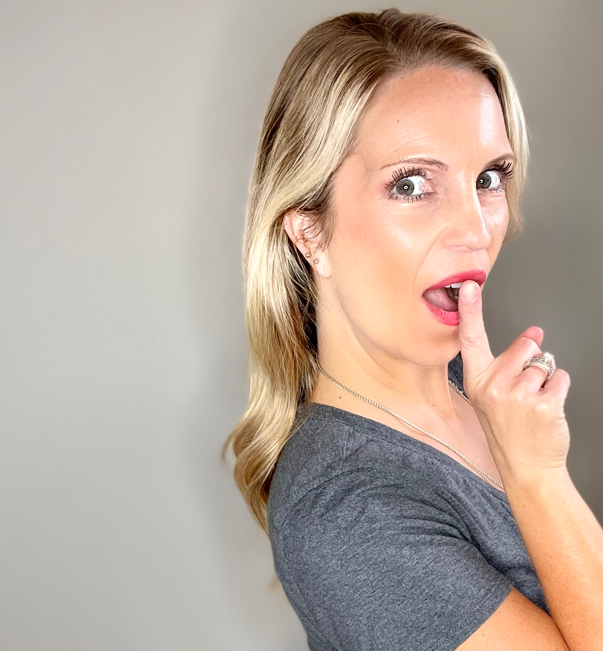 Woman with blonde hair and a gray shirt pointing to her lips against a plain background